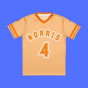Op de afbeelding: Een lichtoranje shirt met het woord "NORRIS" in witte blokletters en het nummer "4" in wit met een oranje omlijning. Het shirt heeft oranje strepen op de mouwen en een V-hals.