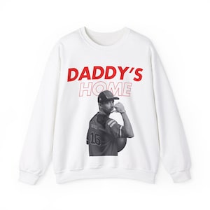 May include: White sweatshirt with red text that reads "DADDY'S HOME". A black and white photo of a man wearing a baseball cap and jersey is printed on the front of the sweatshirt.