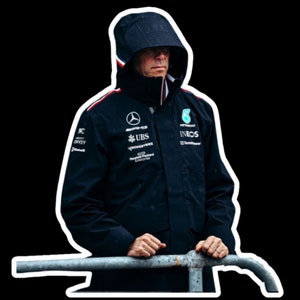 May include: A man wearing a black hooded jacket with various logos, including "INEOS", "UBS", and "Mercedes-AMG", standing by a metal railing.