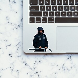 May include: A sticker of a person wearing a black hooded jacket with a white and blue logo on the chest. The person is standing in front of a metal fence.
