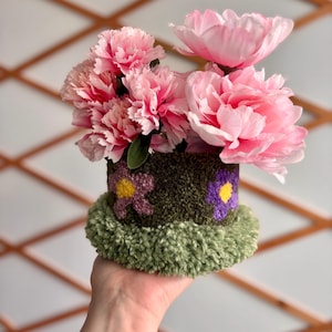 Może przedstawiać: Ręcznie robiona kompozycja kwiatowa z różowymi piwoniami w dekoracyjnym pojemniku. Pojemnik jest ciemnozielony z haftowanymi fioletowymi i żółtymi kwiatami oraz zieloną, puszystą podstawą. Kompozycja jest trzymana przez osobę.