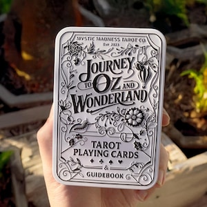 Pode incluir: Uma caixa de cartas de tar&ocirc; branca e prateada com o texto "Journey to Oz and Wonderland" e "Tarot Playing Cards & Guidebook". A caixa tem ilustra&ccedil;&otilde;es florais e caprichosas. A caixa &eacute; segurada em uma m&atilde;o.