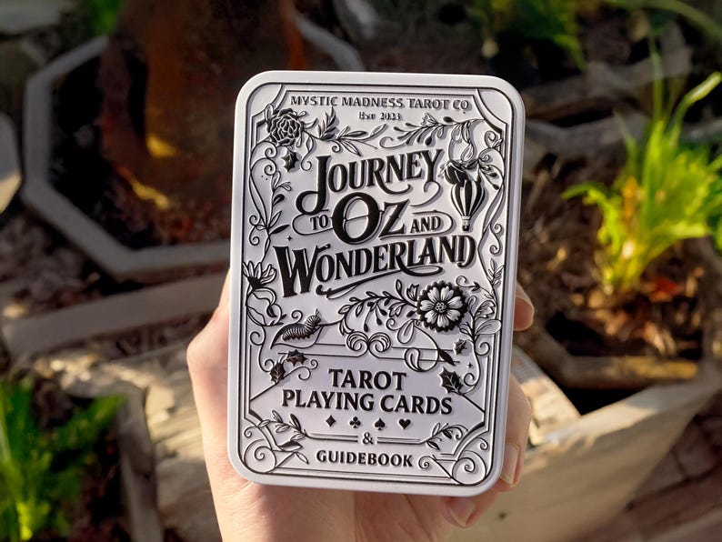 Pode incluir: Uma caixa de cartas de tar&ocirc; branca e prateada com o texto "Journey to Oz and Wonderland" e "Tarot Playing Cards & Guidebook". A caixa tem ilustra&ccedil;&otilde;es florais e caprichosas. A caixa &eacute; segurada em uma m&atilde;o.