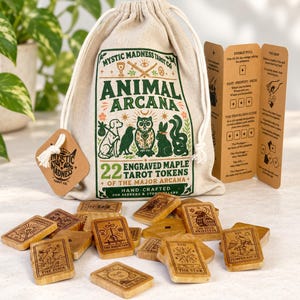 Pode incluir: Um saco de cordão bege com o texto Mystic Madness Tarot Co Animal Arcana 22 Engraved Maple Tarot Tokens of the Major Arcana, acompanhado por um cartão de instruções dobrado e vinte e duas fichas de madeira retangulares gravadas.