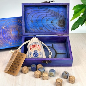 Pode incluir: Uma caixa de madeira roxa com tampa articulada e um design do sistema solar gravado a laser. Dentro, há um saco de cordão creme com a etiqueta ZODIAC DIVINATION DICE, rodeado por pequenos dados de madeira com símbolos.