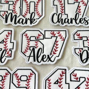 May include: Close-up of several white baseball-themed patches with black outlines. Each patch features a number and a name in black script, with red stitching resembling baseball seams. The patches are for personalization.