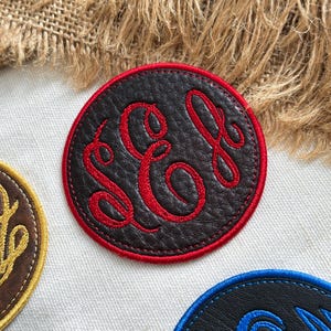 May include: A circular black faux leather patch featuring a red embroidered monogram with the letters S, C, and L in a cursive script. The patch has a thick red stitched border and a textured surface, positioned on a light fabric background.