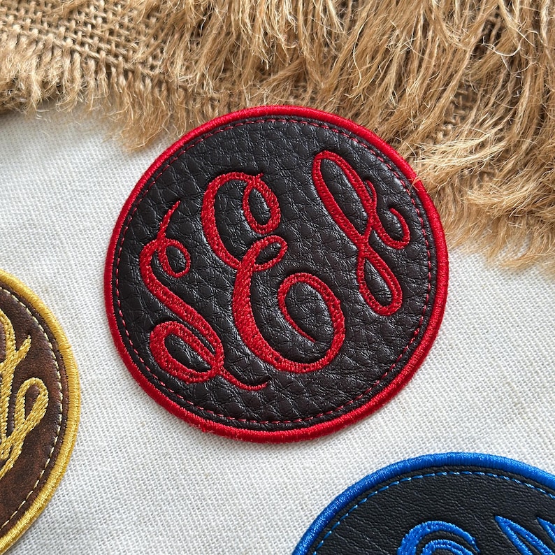 May include: A circular black faux leather patch featuring a red embroidered monogram with the letters S, C, and L in a cursive script. The patch has a thick red stitched border and a textured surface, positioned on a light fabric background.