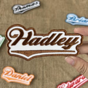 May include: Embroidered patch with the name "Hadley" in brown thread on a white background. The patch has a brown border and is surrounded by other name patches. The patches are likely for clothing or accessories.