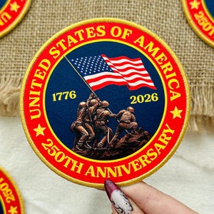 May include: Circular patch with a red border and the text "UNITED STATES OF AMERICA" and "250TH ANNIVERSARY". The patch features a blue center with the American flag and a depiction of soldiers raising the flag. The years 1776 and 2026 are also displayed.