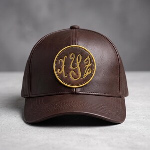 May include: A dark brown baseball cap featuring a circular patch on the front. The patch has a yellow border and contains the embroidered letters XYZ in a cursive script. The cap is constructed with a smooth, leather-like material and stitching.