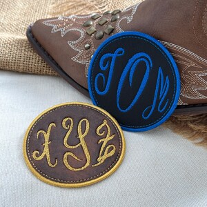 May include: Two circular embroidered patches featuring monogrammed letters. One patch is black with blue stitching and the letters TOM, while the other is brown with gold stitching and the letters XYZ. Both patches have a stitched border.