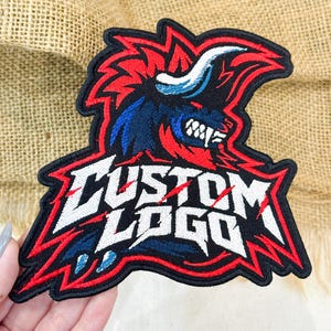 May include: An embroidered patch featuring a stylized dark blue and red bull head with white horns and teeth. The text CUSTOM LOGO is stitched in white with a jagged, distressed font style across the lower portion of the patch, outlined in black.
