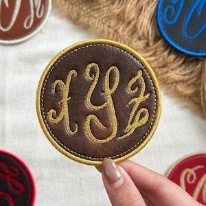 May include: A circular brown leather patch featuring an embroidered gold monogram with the letters X, Y, and Z in a cursive script. The patch has a thick gold stitched border and a smooth texture, showcasing detailed needlework on the surface.