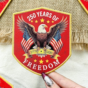 May include: A red patch with a gold border, featuring an eagle with outstretched wings holding American flags. The text reads "250 YEARS OF FREEDOM" with the years 1776 and 2026. Stars and a banner complete the design.