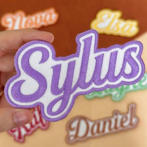 May include: A close-up of a purple and white embroidered patch with the name "Sylus" in a cursive font. Other name patches are visible in the background, including "Nova", "Elsa", "Daniel", and "Sail". The patches are in various colors and fonts.