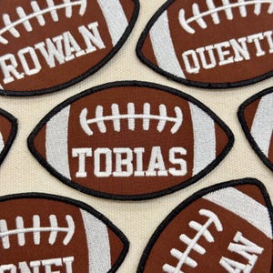 May include: Close-up of several brown football-shaped patches with white stitching and black borders. Each patch has a name embroidered in white. The names include "ROWAN", "QUENTIN", "TOBIAS", and "DANIEL".
