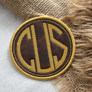 May include: A circular patch featuring a dark brown faux leather base with a gold embroidered border and the gold embroidered monogram letters C, U, and S centered in a stylized font. The patch has a stitched edge and a smooth texture.