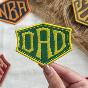 May include: A hexagonal embroidered patch featuring the word DAD in bold yellow lettering against a dark green background. The patch has a thick yellow stitched border and a textured surface, with other similar patches visible in the background.