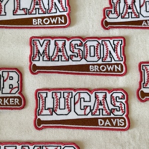 May include: Close-up of several baseball-themed name patches. Each patch features a name in a bold, baseball-style font with red stitching, a baseball bat, and a red border. The names include "MASON", "LUCAS", and others.