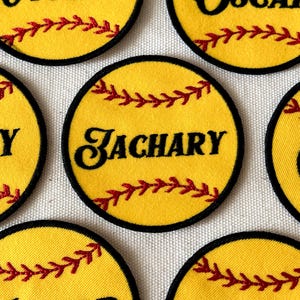 May include: Circular yellow patches resembling softballs, with black edging and red stitching. One patch features the name "ZACHARY" in black script. The patches are designed to look like softballs.