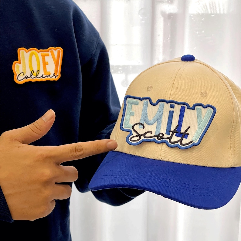 Puede incluir: Gorra de b&eacute;isbol beige con visera azul y bot&oacute;n azul. La gorra presenta el nombre "Emily Scott" bordado en azul y celeste. Tambi&eacute;n se ve una sudadera azul marino con el nombre "Joey Collins" bordado en naranja y amarillo.