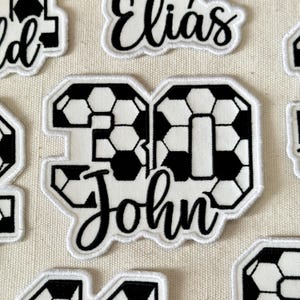 May include: White embroidered patch with the number "30" in a soccer ball pattern, and the name "John" in a cursive font. The patch has a white border and is surrounded by other patches.