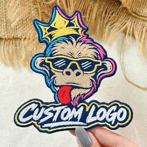 May include: An embroidered patch featuring a monkey wearing yellow sunglasses and a yellow crown, with a red tongue sticking out. Below the monkey, the text CUSTOM LOGO is stitched in white letters with a yellow outline on a dark blue background.