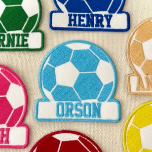May include: Embroidered soccer ball patches in various colors, including blue, green, pink, and red. Each patch features a soccer ball design with a name embroidered on a white banner below. The name "Orson" is visible on a light blue patch.