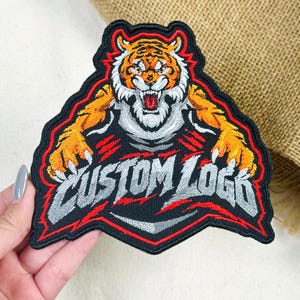 May include: An embroidered patch featuring a snarling tiger with orange, black, and white fur. The tiger has its claws extended over the text CUSTOM LOGO, which is stitched in silver-grey thread. The entire design is outlined in black and red.