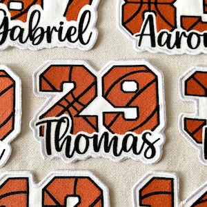 May include: Close-up of several embroidered patches with a basketball theme. The patches feature orange and black basketball designs with white outlines. One patch displays the number 29 and the name "Thomas" in black script.