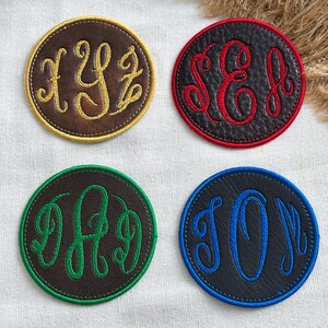 May include: Four circular leather patches with embroidered monograms. The top left is brown with gold stitching, top right is black with red stitching, bottom left is brown with green stitching, and bottom right is black with blue stitching.