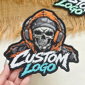 May include: An embroidered patch featuring a skull wearing a beanie and orange headphones with an orange hood. The text CUSTOM LOGO is stitched in white and light blue at the bottom. The patch has a black border and detailed stitching.