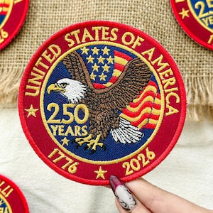 May include: A circular embroidered patch with a patriotic design. It features a bald eagle, the American flag, and the text "UNITED STATES OF AMERICA" and "250 YEARS 1776-2026" in gold lettering on a red and blue background.