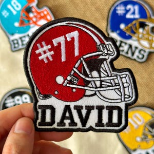 May include: Embroidered patch of a red American football helmet with the number 77 in white. The name "DAVID" is stitched in black letters below the helmet. Other patches with football helmets are visible.