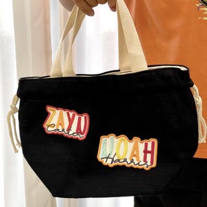 Puede incluir: Bolso tote negro con asas color crema y cierre de cord&oacute;n. El bolso presenta dos parches bordados con los nombres "Zayn Carter" y "Noah Harris" en letras coloridas. El bolso es sostenido por una persona.