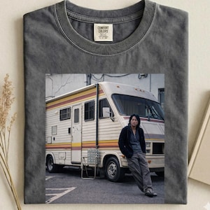 May include: Dark gray Comfort Colors t-shirt featuring a photograph of a person leaning against a vintage RV. The RV has a white body with orange and yellow stripes. The t-shirt has a crew neck and a tag that reads "Comfort Colors."
