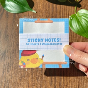 May include: A stack of blue sticky notes with the text "STICKY NOTES!" and "50 sheets | @aiimeecreates". The design includes a cartoon duck wearing a beret and holding a paintbrush. The background is a grid pattern.
