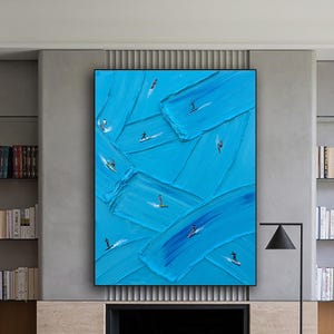 Op de afbeelding: Een groot rechthoekig kunstwerk aan de muur met dikke, getextureerde blauwe olieverfstreken en kleine, abstracte figuren van surfers op golven, tentoongesteld boven een open haard in een kamer met boekenplanken en modern meubilair.