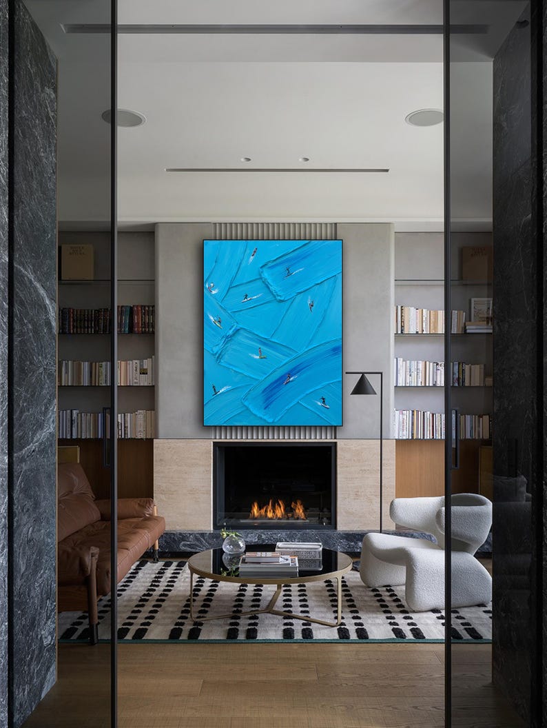 Op de afbeelding: Een groot rechthoekig kunstwerk aan de muur met dikke, getextureerde blauwe olieverfstreken en kleine, abstracte figuren van surfers op golven, tentoongesteld boven een open haard in een kamer met boekenplanken en modern meubilair.