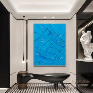Puede incluir: Un cuadro rectangular sobre lienzo con pinceladas gruesas y texturizadas en varios tonos de azul brillante. Peque&ntilde;as figuras abstractas de surfistas sobre tablas est&aacute;n dispersas por la superficie, creando una sensaci&oacute;n de movimiento.