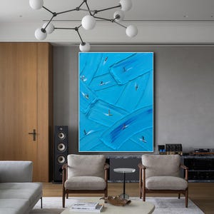 Op de afbeelding: Een groot rechthoekig wandkunstwerk met dikke, getextureerde blauwe verfstreken die over het canvas zijn aangebracht. Kleine, donkere silhouetten van individuen op surfplanken zijn in de blauwe verf geplaatst, wat een gevoel van beweging cre&euml;ert.