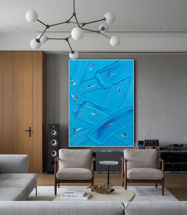 Op de afbeelding: Een groot rechthoekig wandkunstwerk met dikke, getextureerde blauwe verfstreken die over het canvas zijn aangebracht. Kleine, donkere silhouetten van individuen op surfplanken zijn in de blauwe verf geplaatst, wat een gevoel van beweging cre&euml;ert.