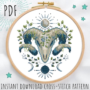 May include: A cross-stitch pattern featuring a ram skull design with blue and green floral accents, set within a wooden embroidery hoop. The design includes a sunburst, crescent moons, and the text "PDF" and "INSTANT DOWNLOAD CROSS-STITCH PATTERN".