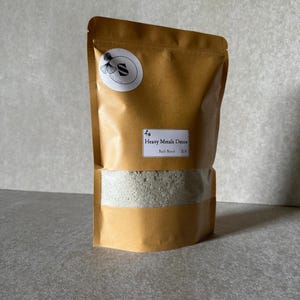 Pode incluir: Um saco de papel castanho com um rótulo branco que diz "Heavy Metals Detox Bath Blend 2.1lb". O saco está cheio de um pó cinzento claro.