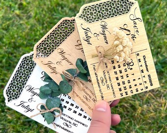 Puede incluir: Tres tarjetas de madera para guardar la fecha con intrincados diseños cortados con láser. Las tarjetas muestran el texto "Save the Date" y los nombres "Wagner & Juliyt". Las tarjetas están decoradas con flores secas y vegetación.
