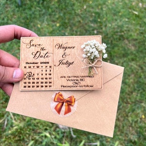May include: A rectangular wooden save the date card featuring a laser-engraved October 2025 calendar with a heart on the 5th, the names Wagner and Juliyt, and a small dried white flower bouquet tied with twine, resting on a brown paper envelope.
