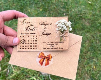 Puede incluir: Una tarjeta de madera rectangular para guardar la fecha con un calendario de octubre de 2025 grabado con láser, un corazón en el día 5, los nombres Wagner y Juliyt, y un pequeño ramo de flores blancas secas atado con cordel sobre un sobre marrón.