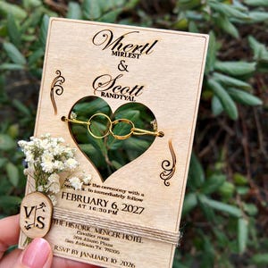 May include: A rectangular wooden wedding invitation card featuring a heart-shaped cutout holding two gold rings. The card displays the names Vhertl Mirlest and Scott Randtyal, the date February 6, 2027, and The Historic Menger Hotel location details.
