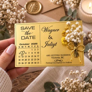 May include: A rectangular gold-colored Save the Date card featuring black text that reads Wagner & Juliyt, October 2025, and Are Getting Married Victoria, BC. A small bouquet of dried white and yellow flowers with a gold ribbon is attached to the right.